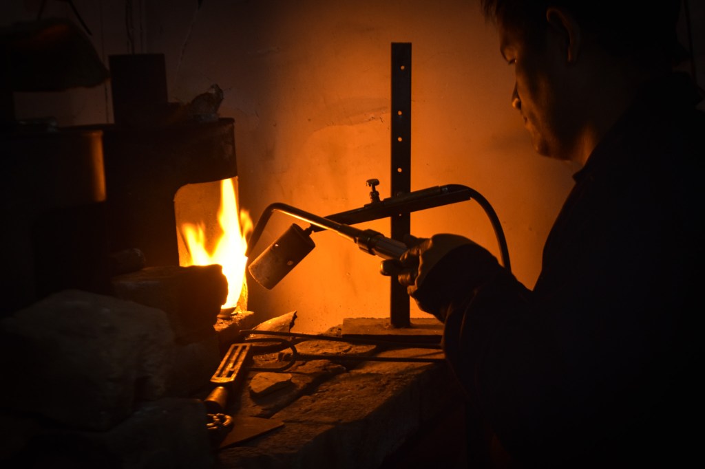 Un joyero trabajando con una antorcha de metal en un entorno oscuro, donde se ve fuego y herramientas de joyería.
